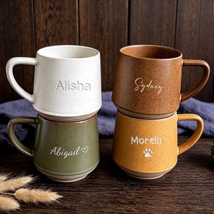 May include: Four ceramic mugs with speckled finishes in white, brown, olive green, and mustard yellow. Each mug features a handle and a personalized name engraved on the front: Alisha, Sydney, Abigail with a heart, and Morelli with a paw print.