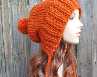 May include: A bright orange knitted beanie with a pom pom on top and long tassels hanging down the back. The beanie is made of a soft, chunky yarn.