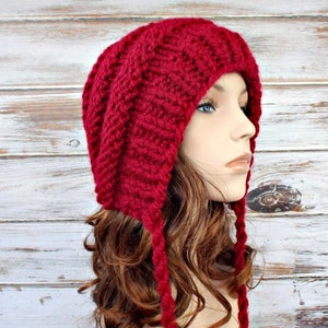 Könnte beinhalten: Eine rote Strickmütze mit langen Bändern. Die Mütze hat eine lockere Passform und ein Zopfmuster.