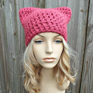 Könnte beinhalten: Eine rosa Häkelmütze mit Katzenohren. Die Mütze sitzt auf einem Mannequin-Kopf mit blonden Haaren.