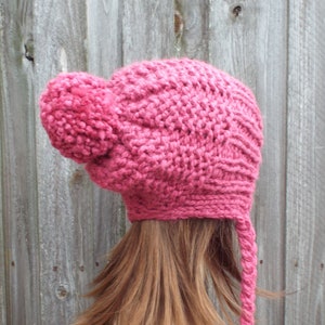 Könnte beinhalten: Eine rosa gestrickte Mütze mit einem großen Bommel oben und einem langen, herabhängenden Band. Die Mütze ist aus weichem, dickem Garn gefertigt.