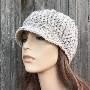 May include: A beige crocheted newsboy cap with a slightly curved brim. The cap is made with a textured stitch and has a speckled appearance.