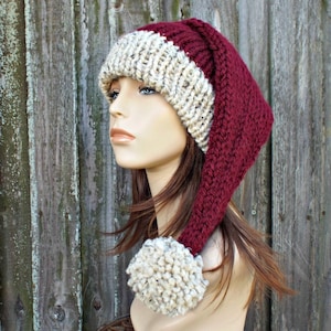 May include: A burgundy and beige knit Santa hat with a long, pointed end and a beige pom-pom. The hat features a thick, textured knit design, perfect for winter wear. The hat is displayed on a mannequin head.