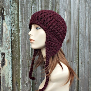 May include: A maroon crocheted hat with long ear flaps. The hat is made with a chunky yarn and has a textured stitch pattern.