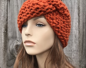 May include: A bright orange knitted headband with a knotted design. The headband is worn on a mannequin with long brown hair.