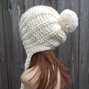 Könnte beinhalten: Eine weiße Strickmütze mit einem Bommel oben und einem langen Zopf. Die Mütze ist aus einem dicken Garn gestrickt und hat ein einfaches Strickmuster.