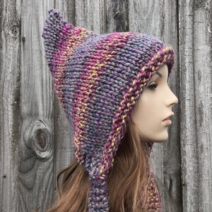 Puede incluir: Un gorro de punto morado, rosa y amarillo con una solapa de oreja larga, puntiaguda y caída. El gorro está hecho con un hilo grueso y tiene un ajuste holgado y caído.