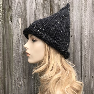 Könnte beinhalten: Eine schwarze gestrickte Hexenmütze mit einer spitzen Krone und einer weißen, gefleckten Textur. Die Mütze wird von einer Puppe mit langen blonden Haaren getragen.