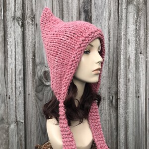 Könnte beinhalten: Eine rosa gestrickte Mütze mit langen Bändern. Die Mütze hat eine spitze Spitze und einen breiten Rand. Die Bänder sind lang genug, um als Schal getragen zu werden.