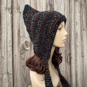 Puede incluir: Un gorro de punto negro, marrón y azul con largas orejeras. El gorro está hecho con un hilo grueso y tiene un ajuste holgado y desgarbado.