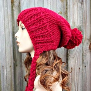 Könnte beinhalten: Eine leuchtend rote, handgestrickte Mütze mit einem langen Zopf und einem großen Bommel. Die Mütze wird auf einem Schaufensterkopf präsentiert und zeigt das strukturierte Strickmuster und das einzigartige Design. Die Ohrenklappen der Mütze gehen in einen langen Zopf über.