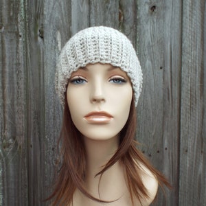 May include: A light beige, ribbed crochet beanie hat. The hat is made of a soft, textured yarn and has a close-fitting design. The hat is shown on a mannequin head with long brown hair and blue eyes. The background is a weathered wooden fence.