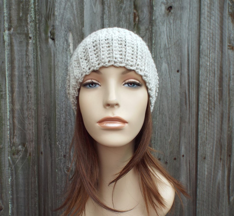 May include: A light beige, ribbed crochet beanie hat. The hat is made of a soft, textured yarn and has a close-fitting design. The hat is shown on a mannequin head with long brown hair and blue eyes. The background is a weathered wooden fence.