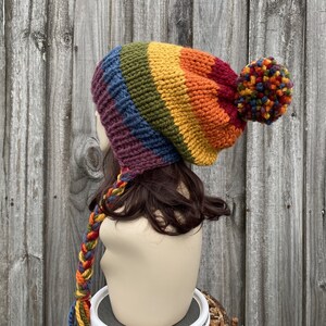 Pu&ograve; includere: Un berretto di lana a righe arcobaleno con un grande pon pon in cima. Il berretto ha un lungo cinturino intrecciato che pende dalla parte posteriore.