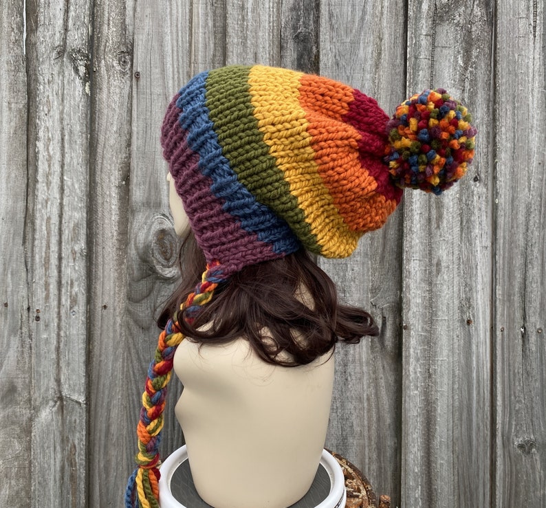 Pu&ograve; includere: Un berretto di lana a righe arcobaleno con un grande pon pon in cima. Il berretto ha un lungo cinturino intrecciato che pende dalla parte posteriore.