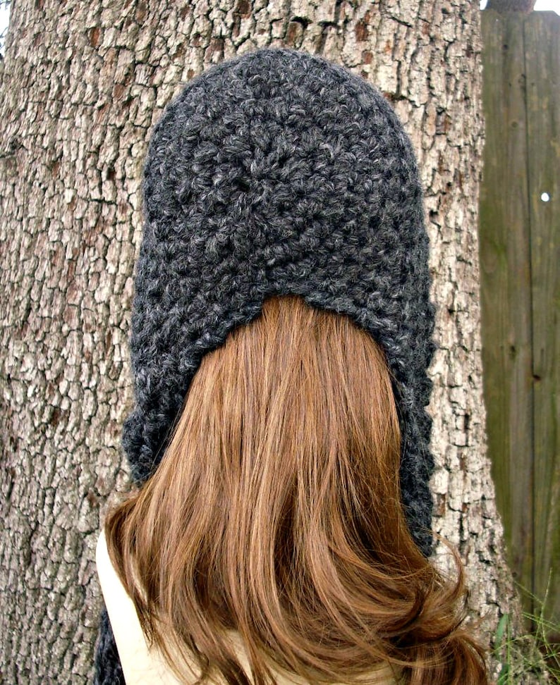 May include: A gray crocheted hat with a long, flowing design. The hat is worn on a person with long brown hair.