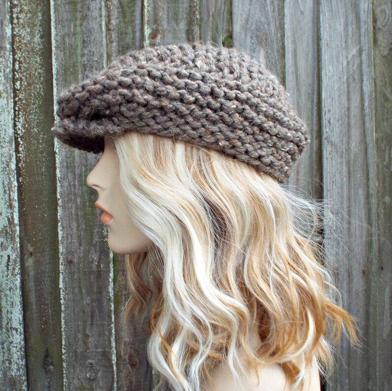 May include: A brown, chunky knit newsboy cap featuring a textured, thick yarn construction with a short, stiff brim. The hat has a rounded, voluminous crown and a speckled appearance created by the multi-tonal brown fibers woven throughout the fabric.