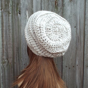 May include: A light beige, handmade crochet hat. The hat is round with a ribbed texture around the brim and a circular pattern on top. The hat is shown from the back, with the wearer's brown hair visible.