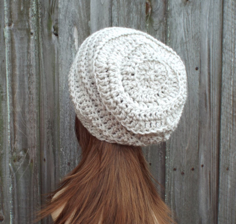 May include: A light beige, handmade crochet hat. The hat is round with a ribbed texture around the brim and a circular pattern on top. The hat is shown from the back, with the wearer's brown hair visible.