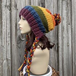 Pu&ograve; includere: Un cappello di lana colorato con un pon pon in cima e lunghi lacci intrecciati. Il cappello &egrave; realizzato in un motivo arcobaleno di rosso, arancione, giallo, verde, blu e viola.