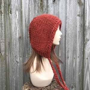 May include: A rust-colored knit hood with long braided ties. The hood is made of a thick, textured yarn and covers the head and neck. The hood is displayed on a mannequin head against a weathered wooden background. The style is reminiscent of a medieval cowl.