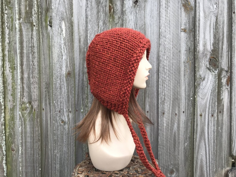May include: A rust-colored knit hood with long braided ties. The hood is made of a thick, textured yarn and covers the head and neck. The hood is displayed on a mannequin head against a weathered wooden background. The style is reminiscent of a medieval cowl.