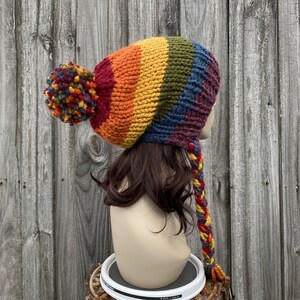 Pu&ograve; includere: Un berretto di lana a righe arcobaleno con un pon pon in cima e un lungo fiocco intrecciato. Il berretto &egrave; fatto di filato di lana ed &egrave; in tonalit&agrave; di rosso, arancione, giallo, verde, blu e viola.