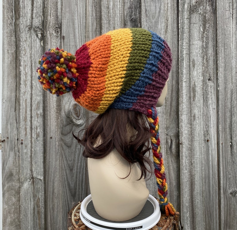 Pu&ograve; includere: Un berretto di lana a righe arcobaleno con un pon pon in cima e un lungo fiocco intrecciato. Il berretto &egrave; fatto di filato di lana ed &egrave; in tonalit&agrave; di rosso, arancione, giallo, verde, blu e viola.
