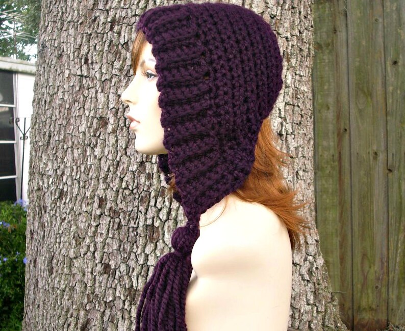 Puede incluir: Un gorro de punto de color morado con una larga bufanda adjunta. El gorro tiene una parte superior puntiaguda y cubre las orejas. La bufanda est&aacute; anudada en la parte delantera.
