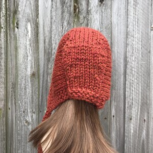 May include: A rust-colored knit hat with ear flaps and a long tie. The hat is made of a thick, textured yarn and is shown against a weathered wooden background. The hat is a warm, cozy accessory.