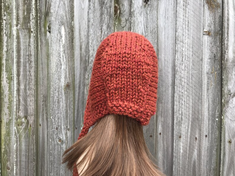 May include: A rust-colored knit hat with ear flaps and a long tie. The hat is made of a thick, textured yarn and is shown against a weathered wooden background. The hat is a warm, cozy accessory.
