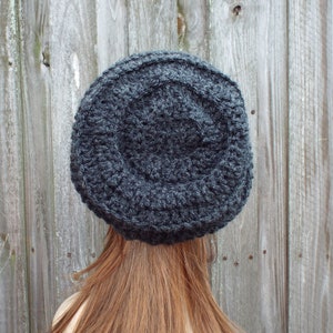 May include: A dark gray, hand-crocheted hat. The hat is round with a textured surface and a slightly raised crown. The hat is made of a thick yarn and appears warm and cozy. The hat is shown from the back, with long auburn hair visible.