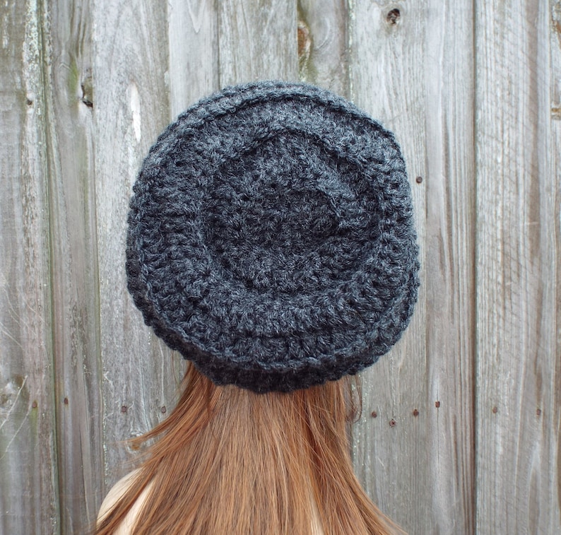 May include: A dark gray, hand-crocheted hat. The hat is round with a textured surface and a slightly raised crown. The hat is made of a thick yarn and appears warm and cozy. The hat is shown from the back, with long auburn hair visible.