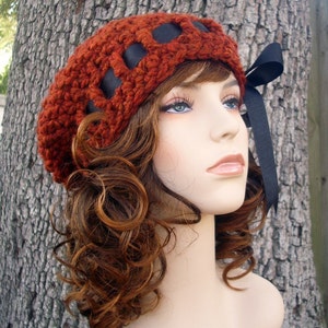 May include: A crocheted beret in a deep red color with black ribbon accents. The beret is worn on a mannequin with long, curly brown hair.