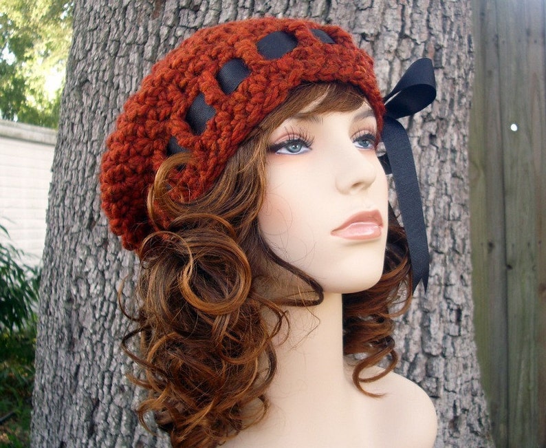 May include: A crocheted beret in a deep red color with black ribbon accents. The beret is worn on a mannequin with long, curly brown hair.