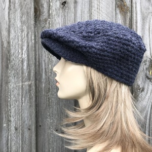 May include: A navy blue, crocheted newsboy cap with a textured pattern. The hat features a rounded crown and a small, stiff brim. The cap is displayed on a mannequin head against a weathered wooden background.