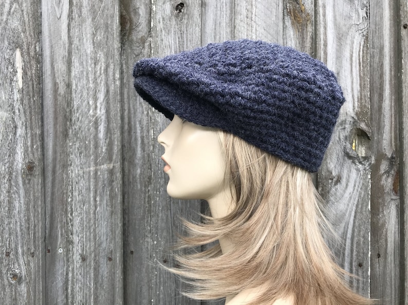 May include: A navy blue, crocheted newsboy cap with a textured pattern. The hat features a rounded crown and a small, stiff brim. The cap is displayed on a mannequin head against a weathered wooden background.