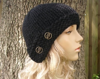 May include: A black knitted cloche hat with two decorative brass buttons. The hat is angled to the side and has a simple, classic design.