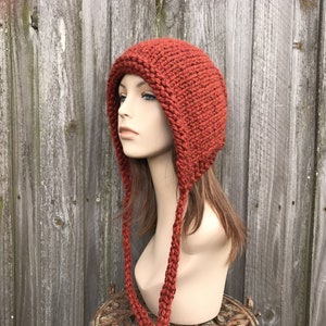 May include: A rust-colored knit hat with ear flaps and long braided ties. The hat is made of thick yarn and has a textured appearance. The hat is displayed on a mannequin head against a weathered wooden background.