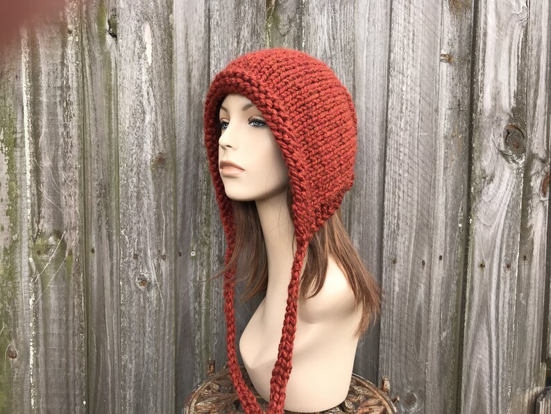 May include: A rust-colored knit hat with ear flaps and long braided ties. The hat is made of thick yarn and has a textured appearance. The hat is displayed on a mannequin head against a weathered wooden background.