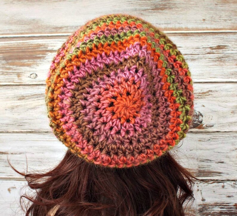 Puede incluir: Un gorro de punto de ganchillo en tonos marrones, rosas, naranjas y verdes. El gorro tiene un dise&ntilde;o circular con un patr&oacute;n de punto texturizado.