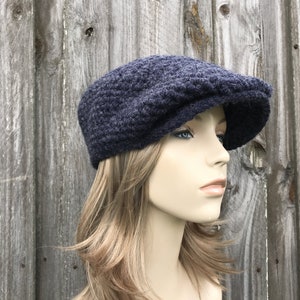 May include: A navy blue, knitted newsboy cap with a textured pattern. The cap has a rounded crown and a short, stiff brim. The hat is displayed on a mannequin with blonde hair against a weathered wooden background.