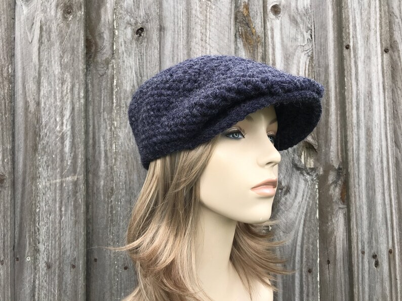 May include: A navy blue, knitted newsboy cap with a textured pattern. The cap has a rounded crown and a short, stiff brim. The hat is displayed on a mannequin with blonde hair against a weathered wooden background.