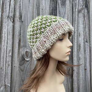 May include: A knitted beanie hat in a green and cream color pattern. The hat is made with a chunky yarn and has a slouchy fit.