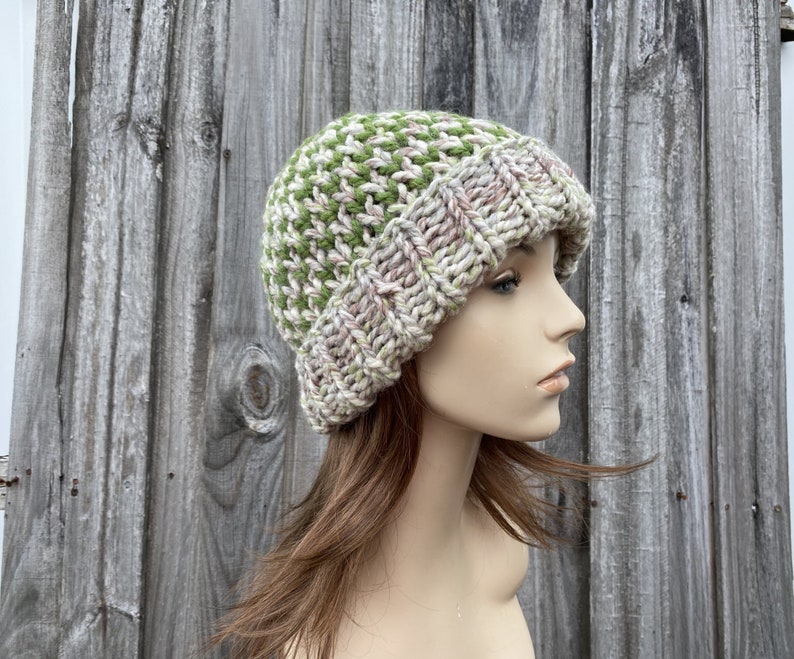 May include: A knitted beanie hat in a green and cream color pattern. The hat is made with a chunky yarn and has a slouchy fit.