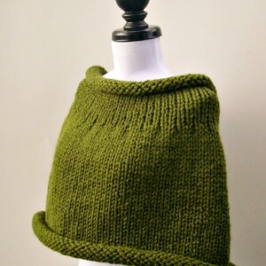 May include: A green knitted cape or shawl with a simple design. The cape is displayed on a white mannequin with a black stand.