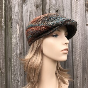 Op de afbeelding: Een gebreide newsboy-pet in bruin, teal en oranje. De pet heeft een platte bovenkant en een gebogen rand.