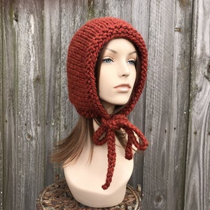 May include: A rust-colored, hand-knitted hood with a rounded top and a tie closure. The hood is displayed on a mannequin head against a weathered wooden fence. The knitted texture is clearly visible.