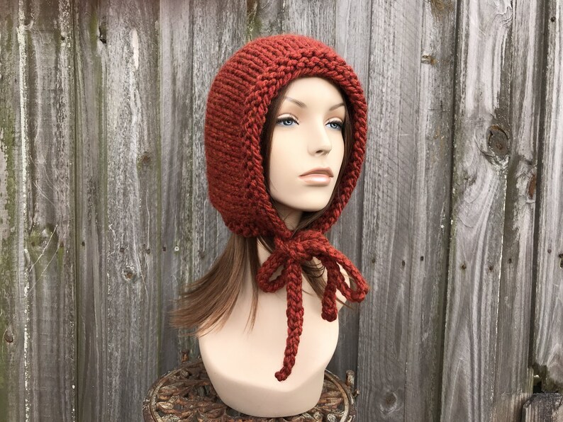 May include: A rust-colored, hand-knitted hood with a rounded top and a tie closure. The hood is displayed on a mannequin head against a weathered wooden fence. The knitted texture is clearly visible.