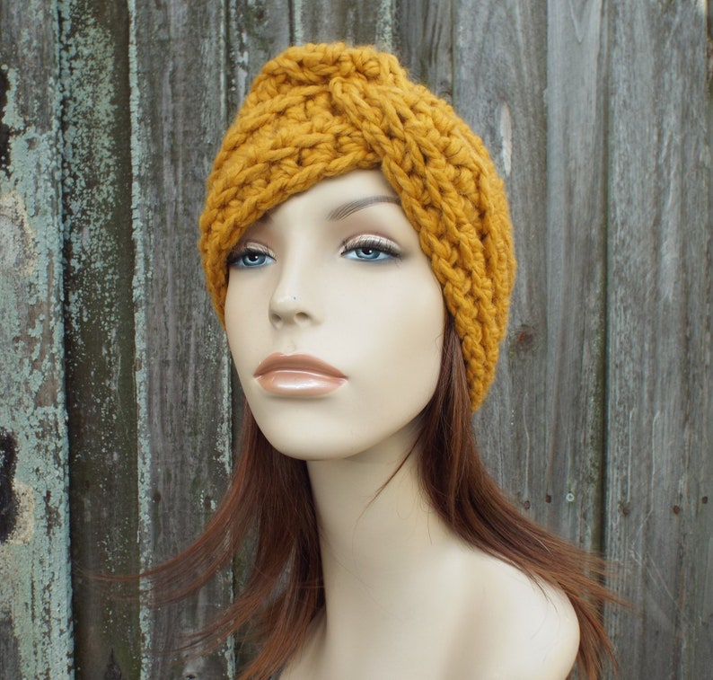 K&ouml;nnte beinhalten: Ein geh&auml;keltes Stirnband in Senfgelb mit einem geknoteten Design. Das Stirnband wird von einer Mannequin mit langen braunen Haaren getragen.